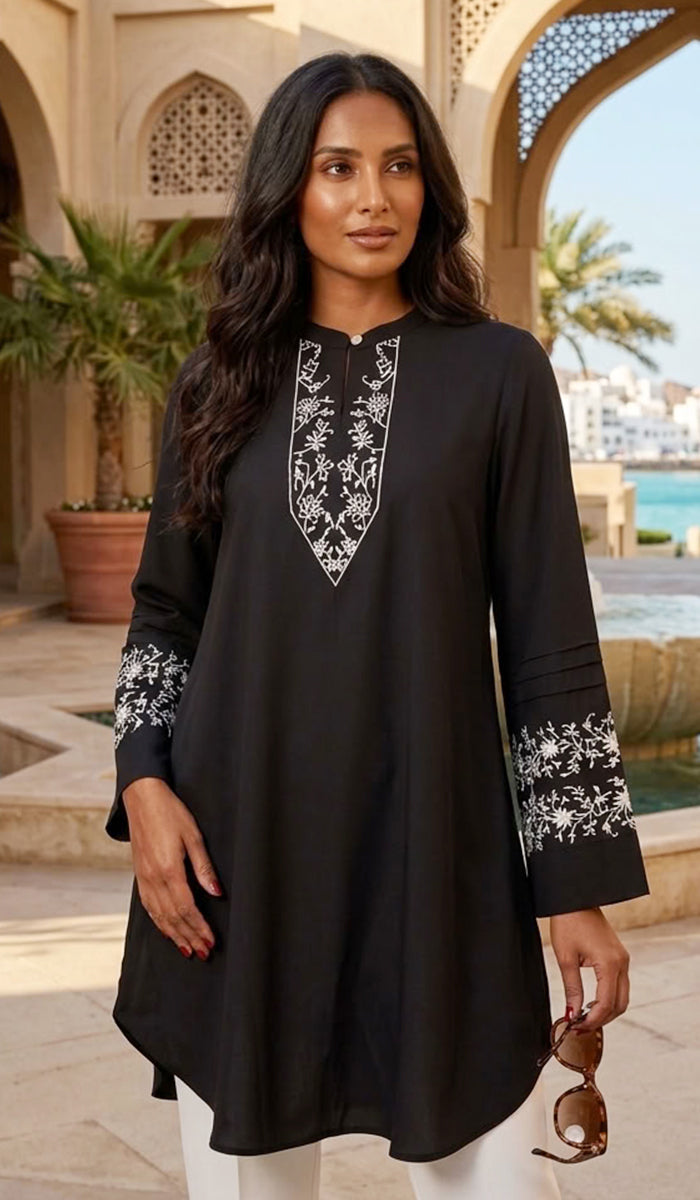 A woman models a black long-sleeve tunic top with white floral embroidery on the neckline and cuffs, set against an outdoor resort background with arches and a pool.