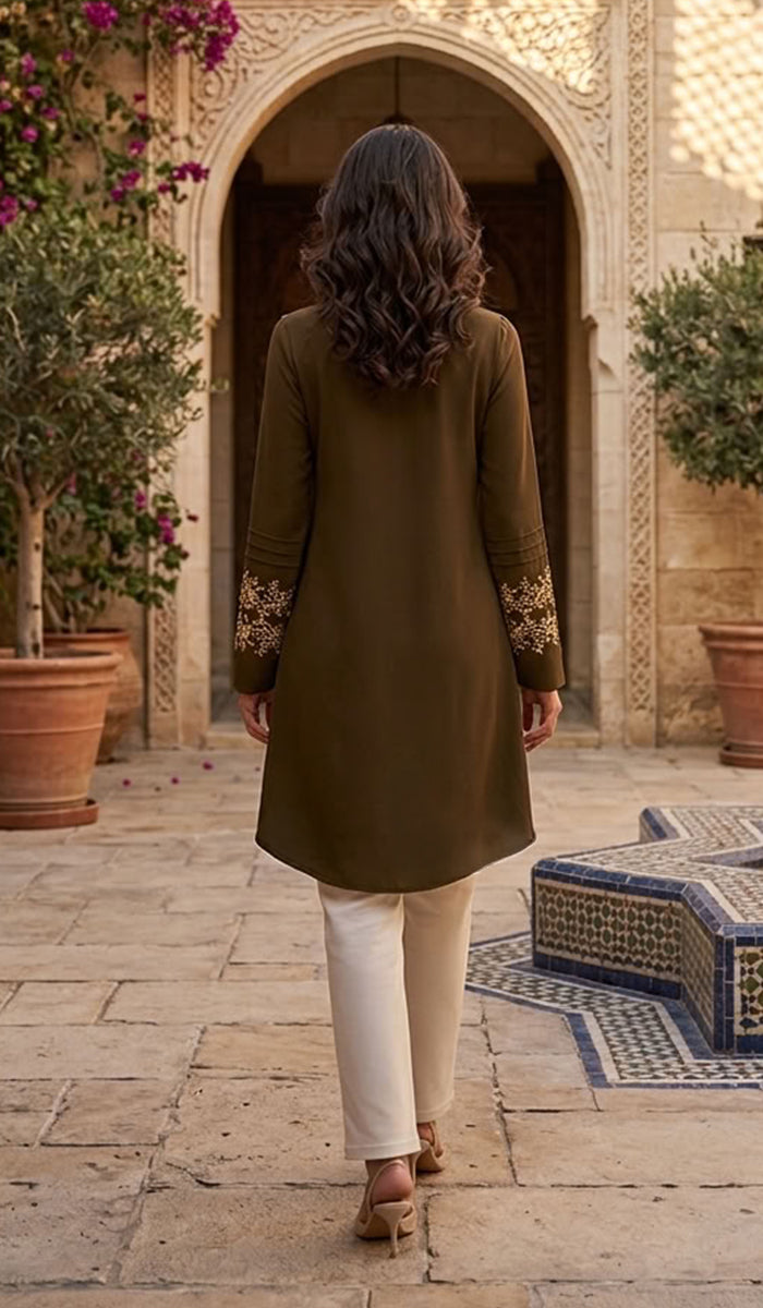 A woman viewed from behind walking through a courtyard wearing an olive green tunic with gold embroidery on the sleeves, paired with cream pants and brown heels.