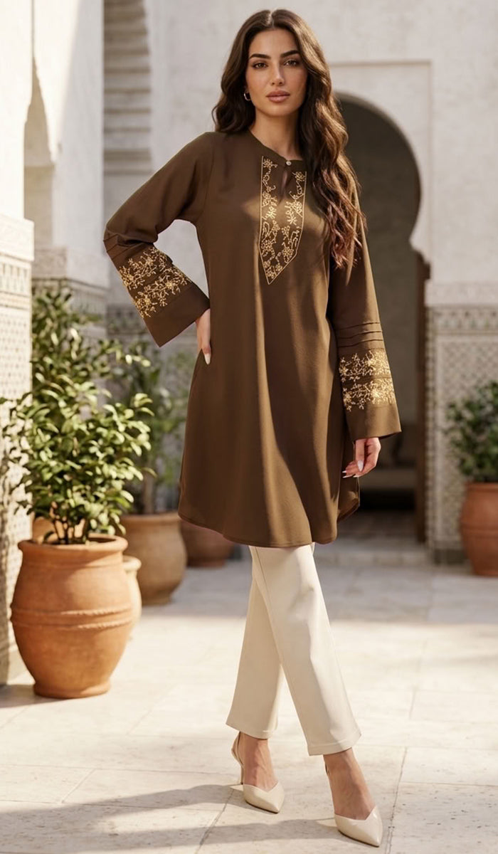 A woman models a brown long-sleeve tunic with gold embroidery on the chest and cuffs, paired with cream pants and beige heels, set against an outdoor courtyard background with potted plants.