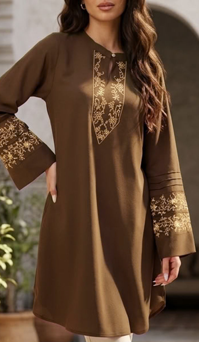 A woman wearing a brown long-sleeve tunic dress with gold floral embroidery on the chest and cuffs, photographed in a softly lit indoor setting.