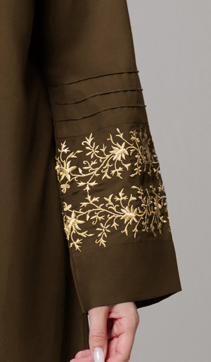 Close-up of a dark brown garment sleeve with gold floral embroidery near the cuff and a hand holding the hem against a plain gray background.