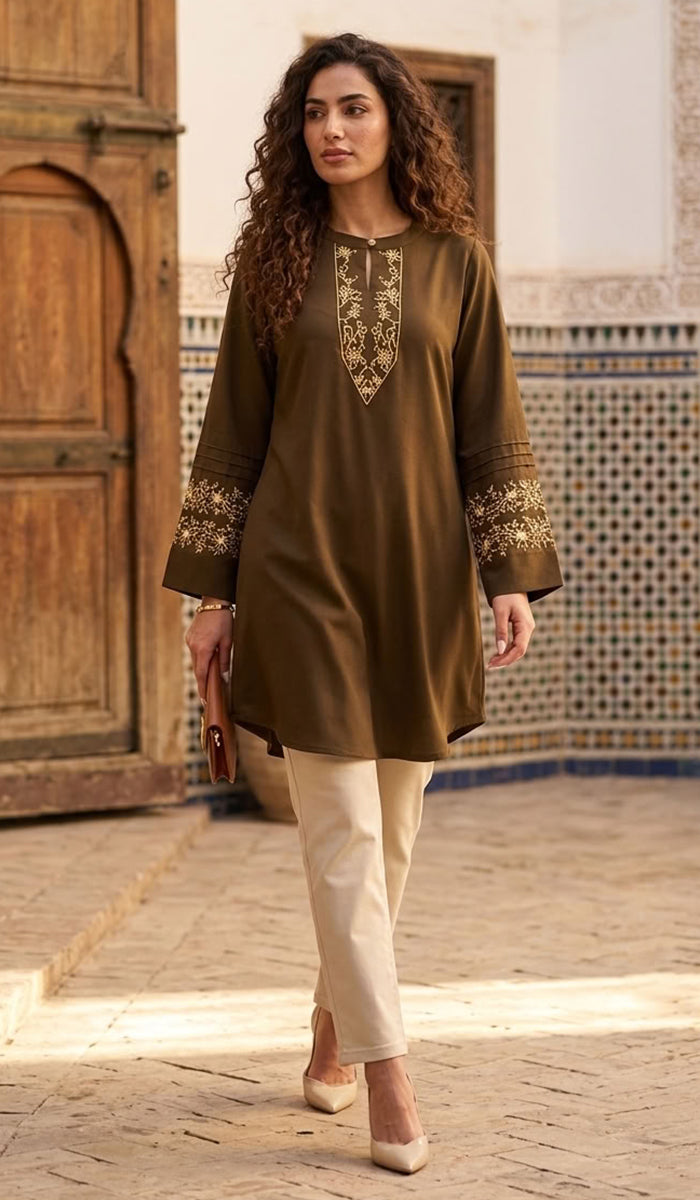 A woman wearing a brown long-sleeve tunic with gold embroidery on the neckline and cuffs, paired with cream pants, walking in a courtyard with a wooden door and tiled wall in the background.