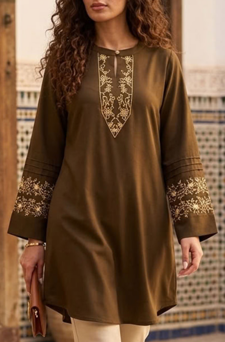 A woman wearing a brown long-sleeve tunic with gold embroidery on the neckline and cuffs, standing in front of a patterned tile wall.