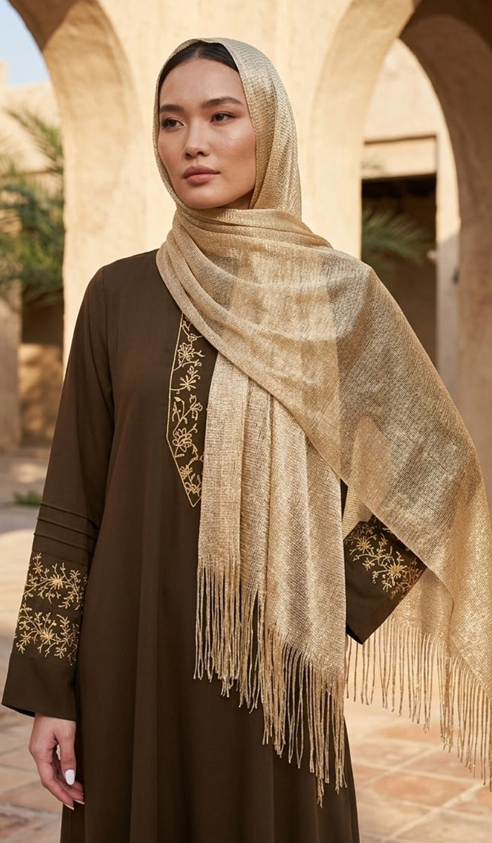 A woman wearing a dark brown abaya with gold embroidery and a shimmering beige hijab, standing in front of a warm-toned stone archway.