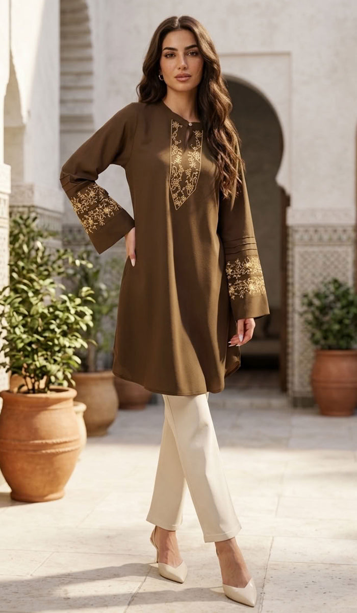 A woman models a brown long-sleeve kurta with gold embroidery on the chest and cuffs, paired with cream pants and beige heels, set against a Moroccan-style courtyard background.