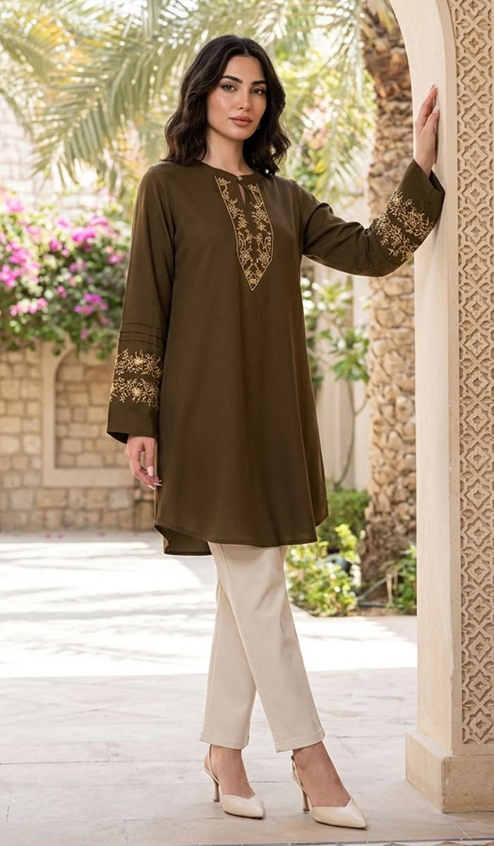 A woman models a long-sleeved olive green tunic with gold embroidery on the neckline and cuffs, paired with cream-colored pants and beige heels, standing in an outdoor courtyard setting.