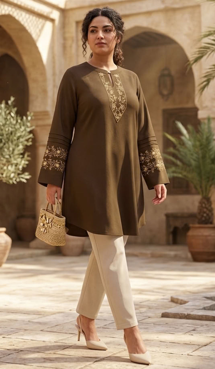 A woman models a dark brown long kurta with gold embroidery on the neckline and cuffs, paired with beige trousers and holding a small woven bag, set against an outdoor courtyard with arches.