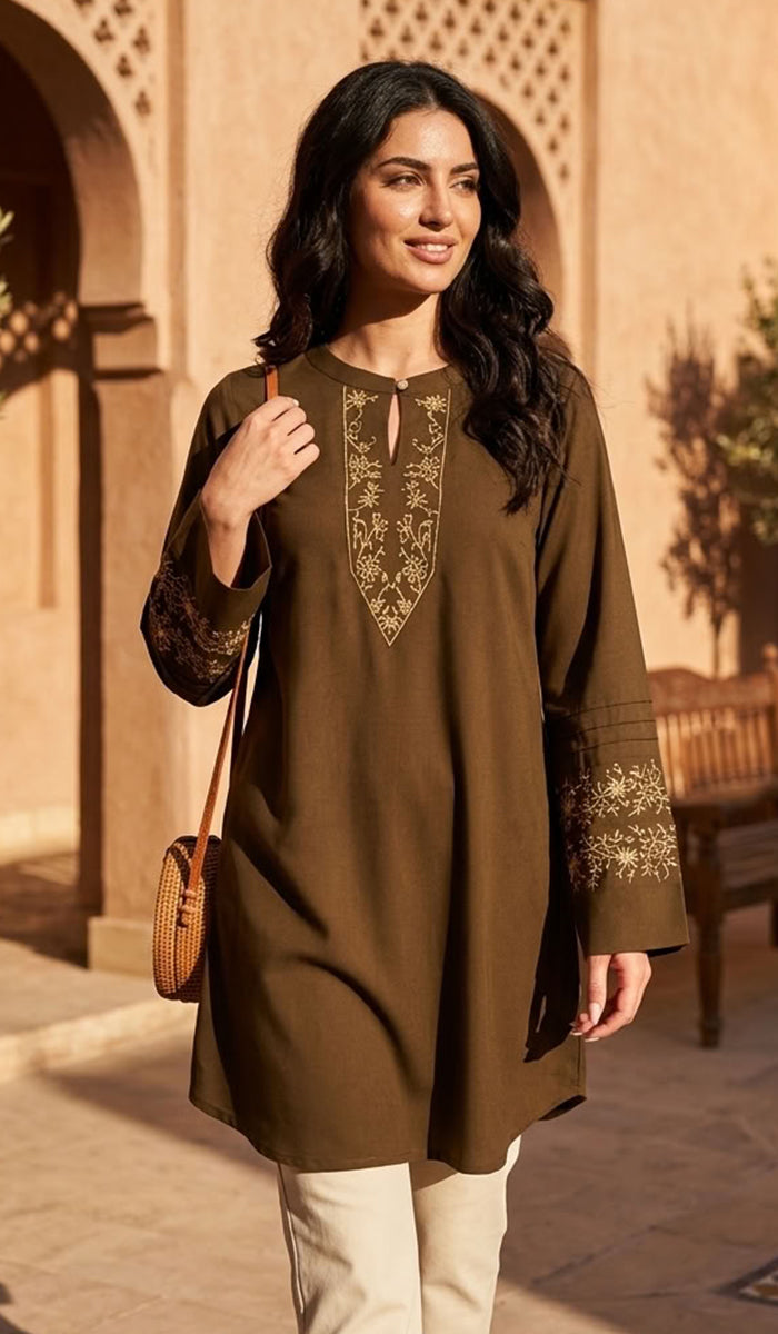 A woman wearing a brown long-sleeve kurta with gold embroidery on the neckline and cuffs, paired with cream pants and carrying a small brown woven bag, standing in front of a warm-toned architectural background.