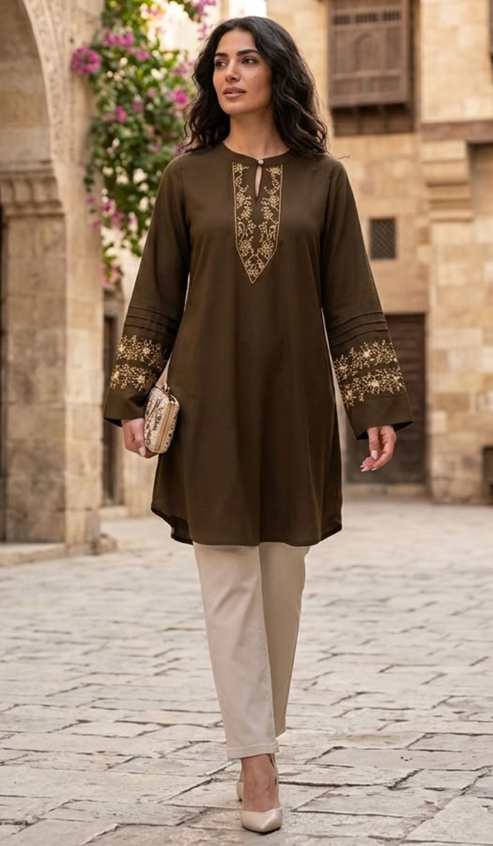 A woman models a dark brown long-sleeve kurta with gold embroidery on the neckline and cuffs, paired with beige trousers, walking on a cobblestone street in front of a stone building.