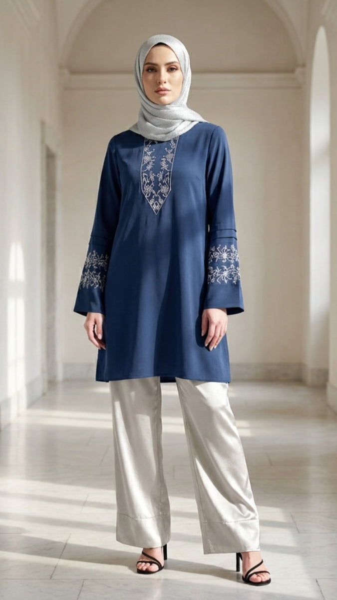 Woman wearing a blue embroidered top and white pants in a marble hallway