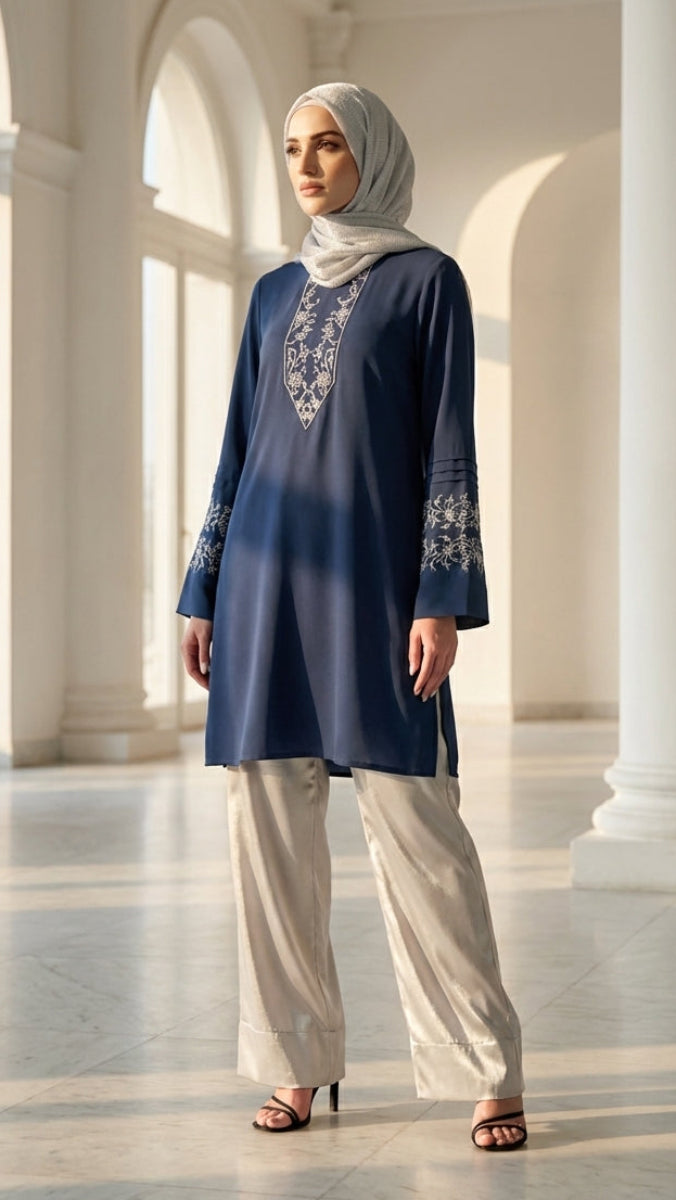 Woman in a blue embroidered top and light pants standing in a bright, architecturally designed room.