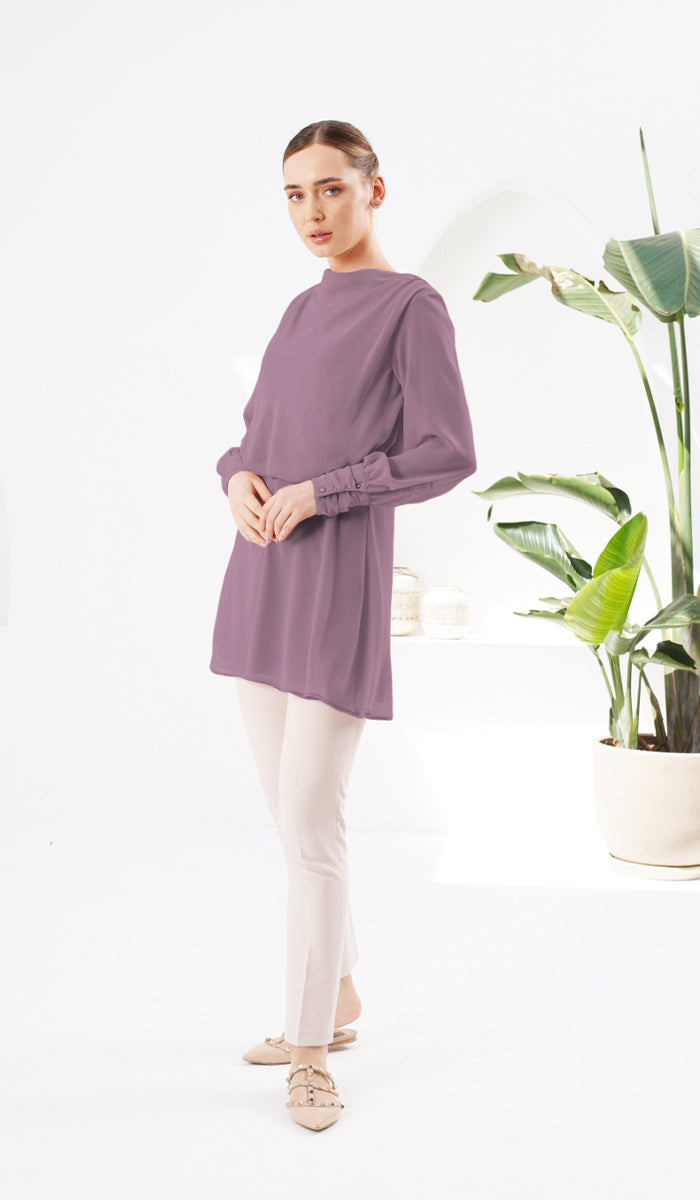Woman next to plant wearing Myra chiffon tunic in Dusty Mauve with white pants and flats. 